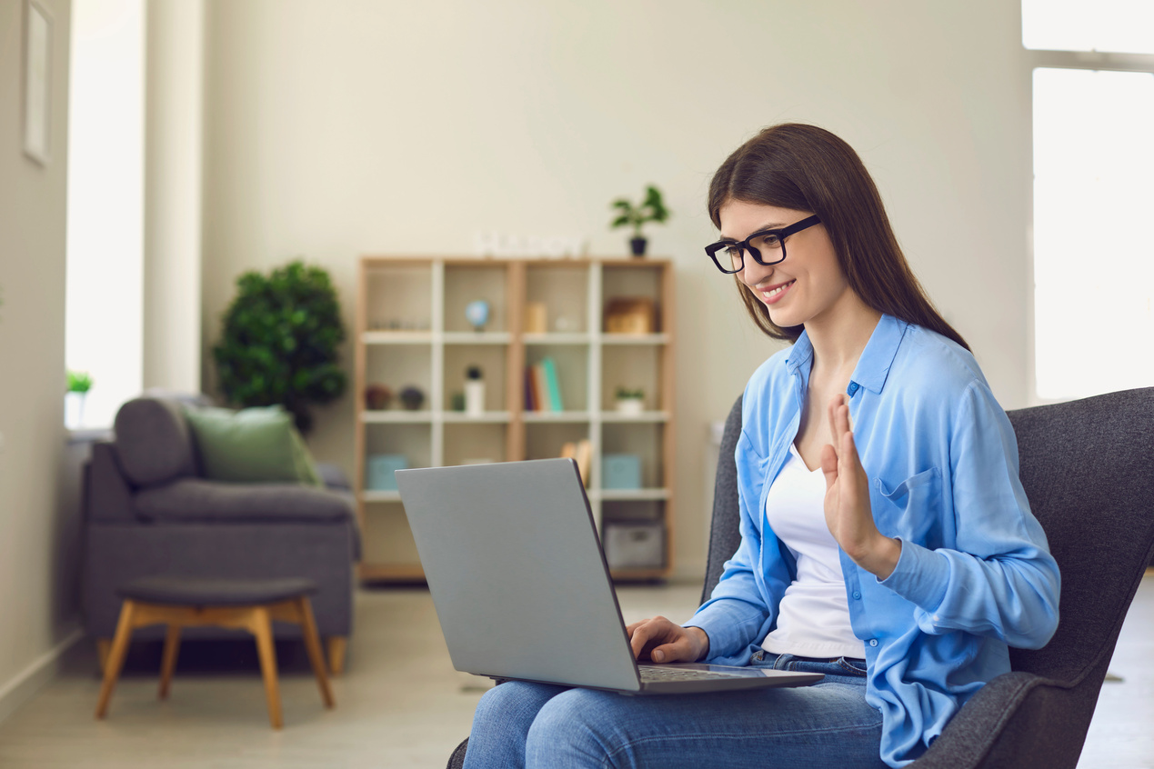 Young Woman in Glasses Making Video Call on Laptop and Waving Hand to Greet Relatives or Colleagues